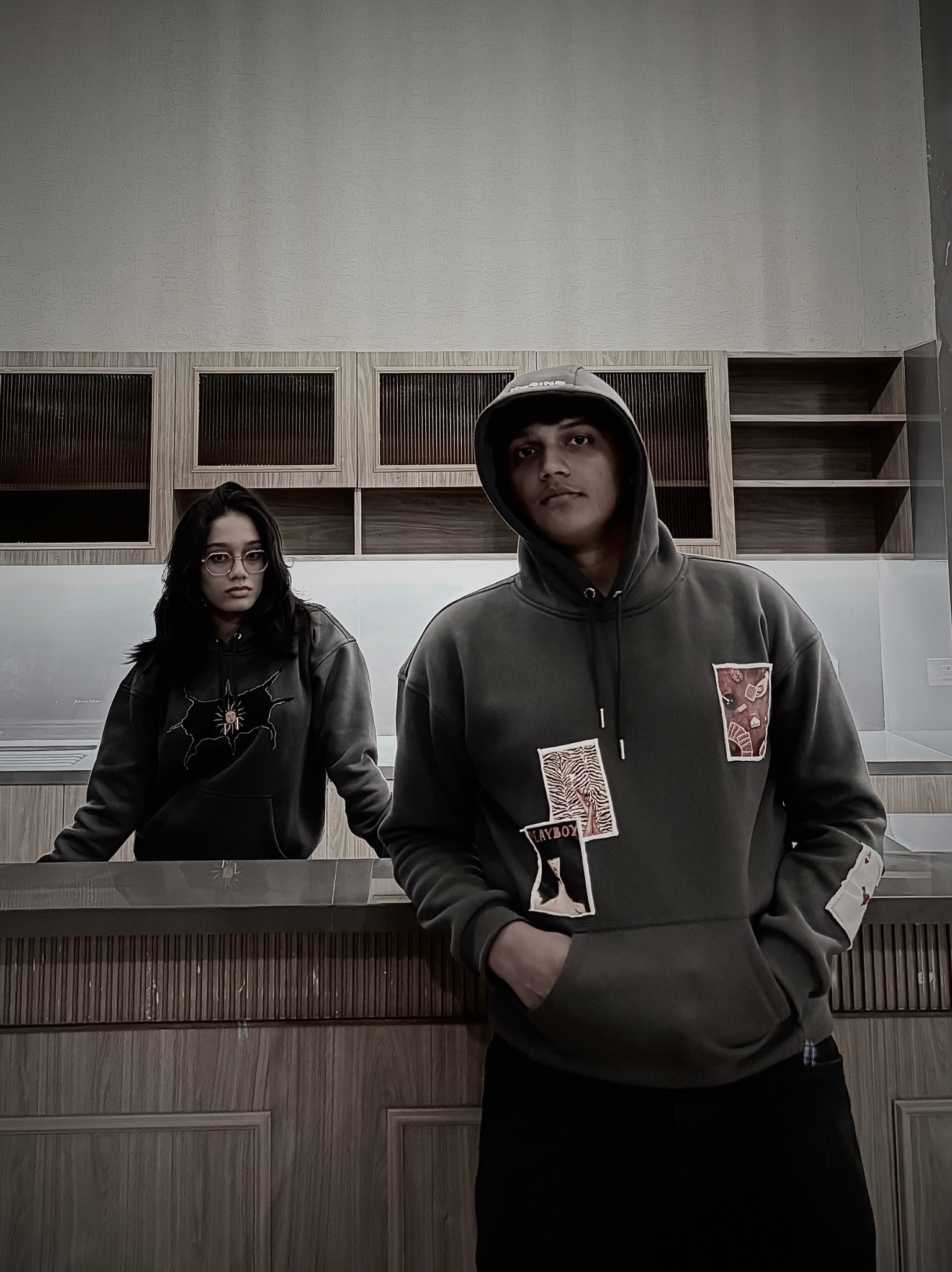 Two people wearing hoodies in a room with wooden shelves.
